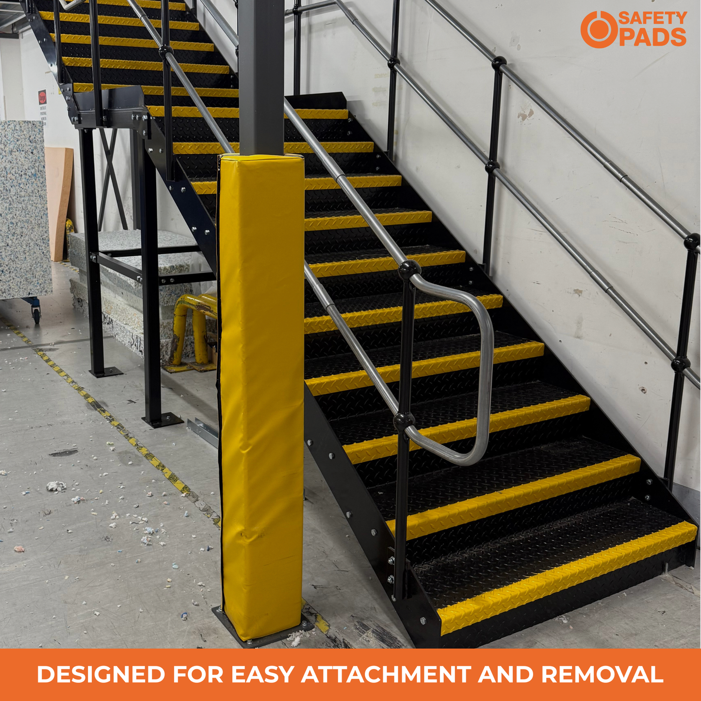 Industrial metal staircase with black steps and yellow safety striping, featuring a yellow protective pad on a post and a silver handrail. The image includes 'SAFETY PADS' branding and an orange banner at the bottom reading 'DESIGNED FOR EASY ATTACHMENT AND REMOVAL'.