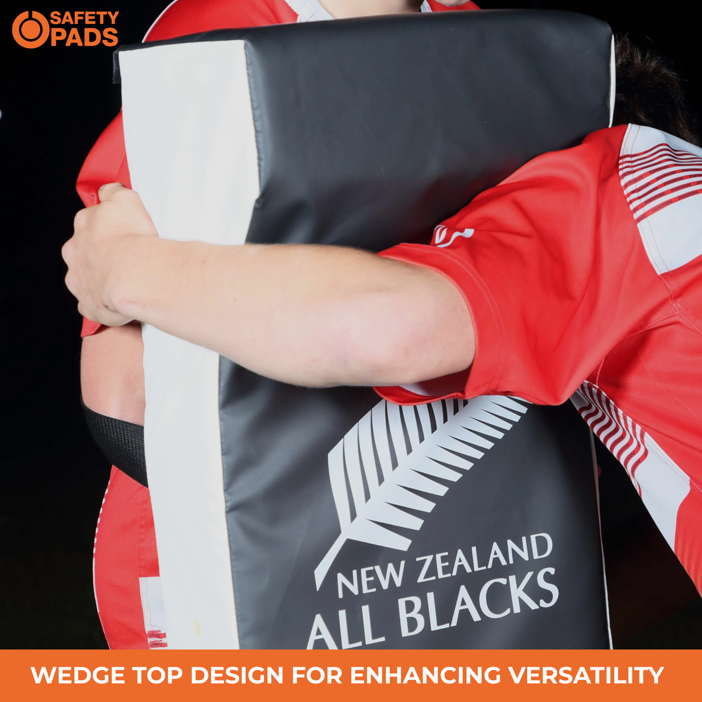 A close-up of a person in a red and white New Zealand All Blacks jersey holding a black and white wedge-shaped protective pad with the New Zealand All Blacks logo. The image includes a 'Safety Pads' logo in the top-left corner and an orange text banner at the bottom.