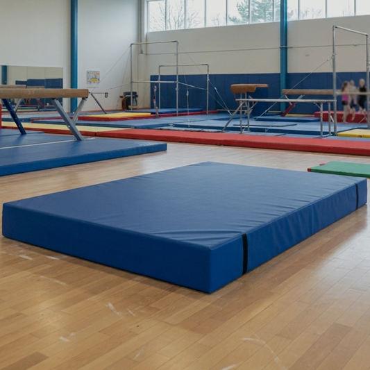 A blue gymnastics floor mat with integrated carry handles on a wooden gym floor, with other gymnastics equipment and a child visible in the background.