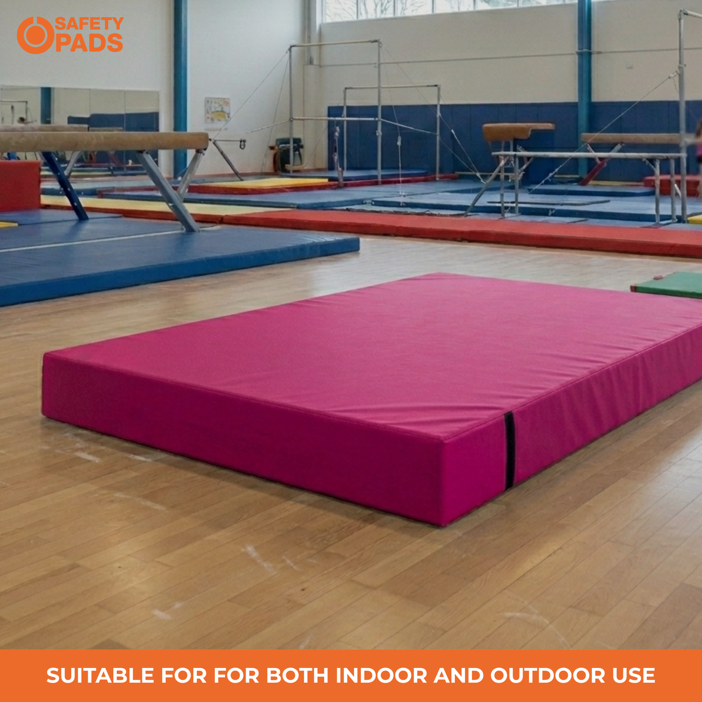 A bright pink gymnastics mat placed on a wooden floor in an indoor gymnastics gym, with balance beams, trampolines, and other mats visible in the background. An orange 'Safety Pads' logo appears in the top-left corner, and an orange banner at the bottom reads 'Suitable for both indoor and outdoor use'.