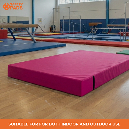 A bright pink gymnastics mat placed on a wooden floor in an indoor gymnastics gym, with balance beams, trampolines, and other mats visible in the background. An orange 'Safety Pads' logo appears in the top-left corner, and an orange banner at the bottom reads 'Suitable for both indoor and outdoor use'.