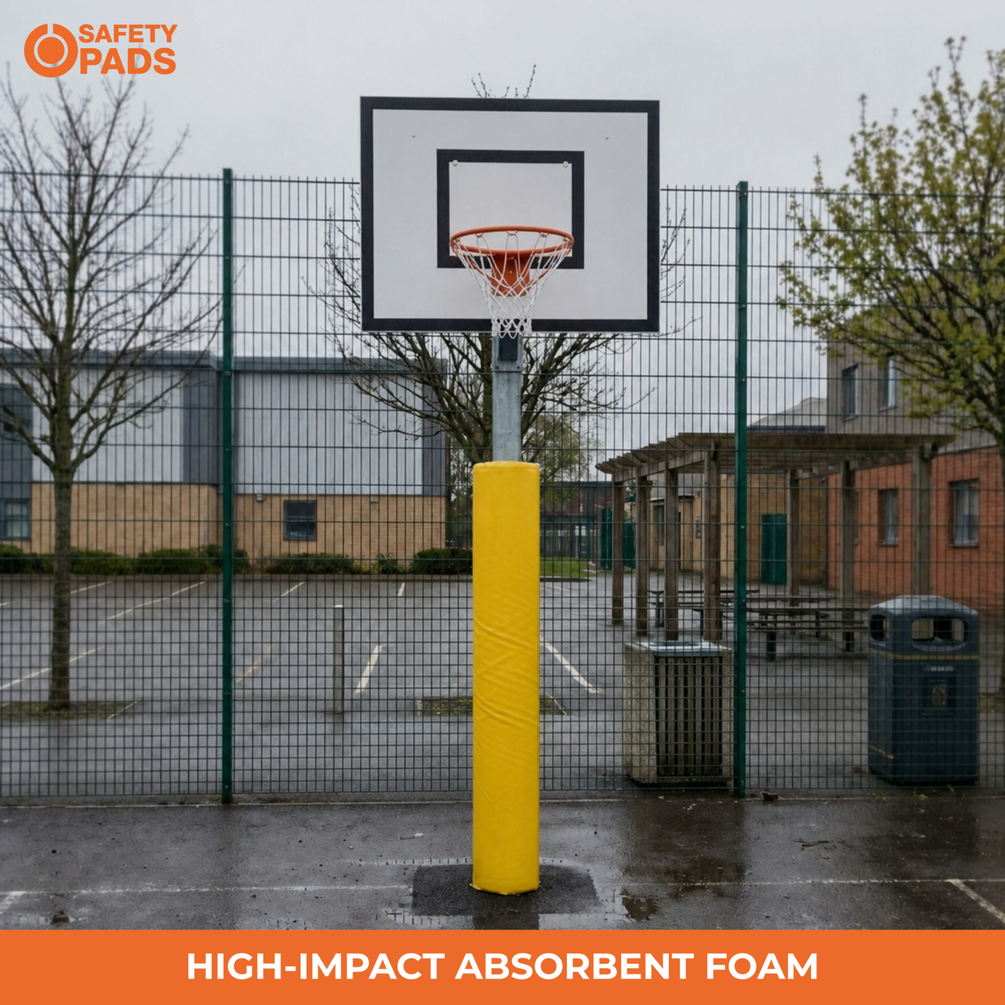 A basketball hoop with a bright yellow high-impact absorbent foam guard in an outdoor court setting. The image includes the 'Safety Pads' brand logo in the top-left corner and an orange banner at the bottom reading 'HIGH-IMPACT ABSORBENT FOAM'.