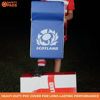 A person outdoors at night holds a blue Scotland rugby pad with the white Scotland rugby logo and text. The person is wearing a red and white sports jersey, white gloves, and black cleats, standing on top of a red and white box.
