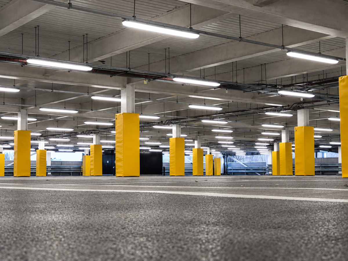 An empty indoor parking structure with concrete pillars, fluorescent lighting, and bright yellow protective bollards on the columns.