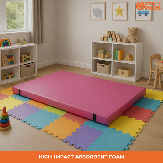 A pink high-impact absorbent foam mattress topper or play mat displayed in a child's room on top of colorful interlocking foam floor mats. The room includes white shelves with toys, a teddy bear, framed wall art, and a window.