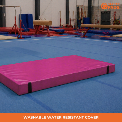 A pink gymnastics landing pad with a black strap closure, placed on a blue gym floor. The background shows other gymnastics equipment in an indoor gym.