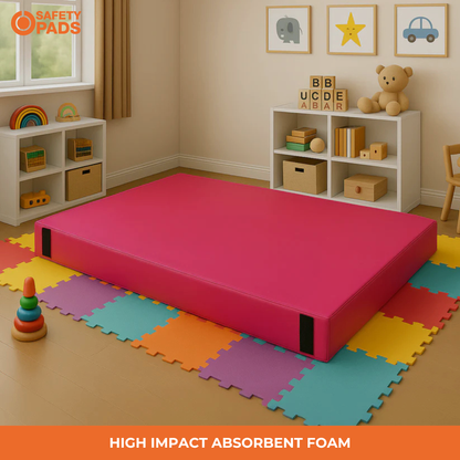 A bright pink safety foam mattress or play mat shown in a styled children's room with colorful interlocking foam mats on the floor, toy shelves, and a window. The image includes the Safety Pads logo and the text 'HIGH IMPACT ABSORBENT FOAM'.