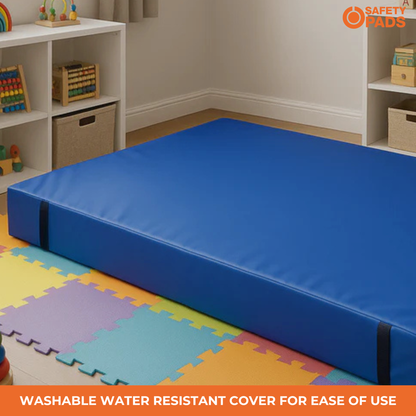 A blue foam safety mat or mattress cover shown in a children's room setting with colorful floor mats, white shelves, and toys. An orange 'SAFETY PADS' logo appears in the top right, and a text banner at the bottom reads 'WASHABLE WATER RESISTANT COVER FOR EASE OF USE'.