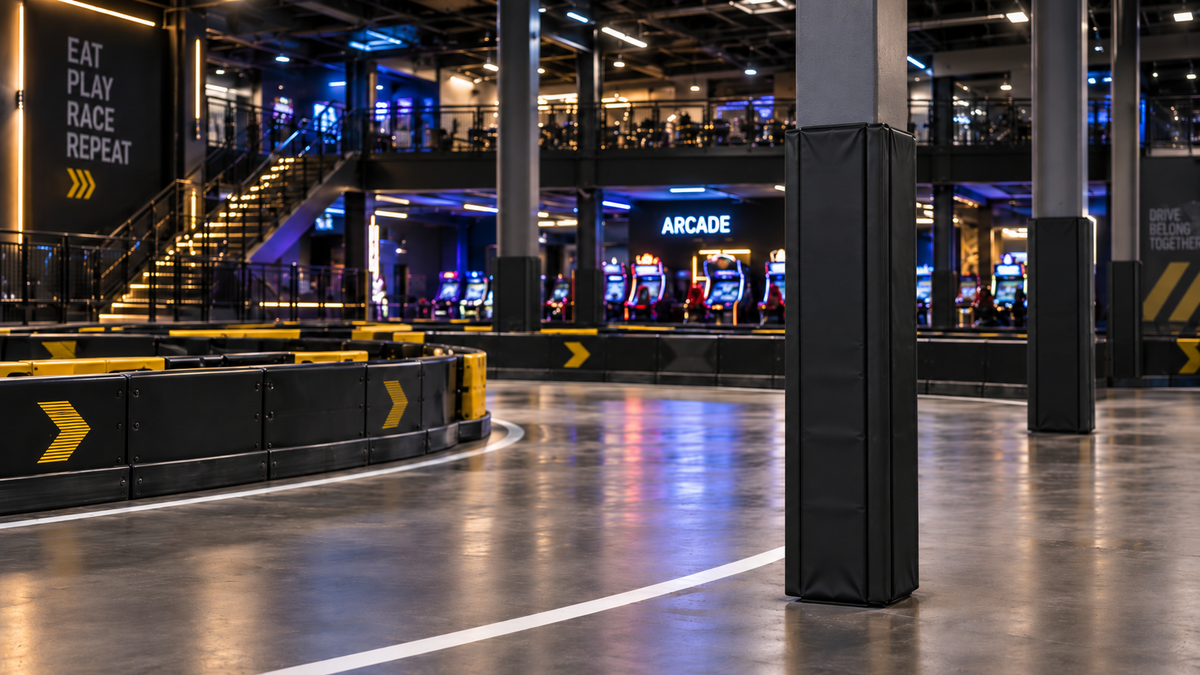 A wide interior view of a modern arcade and gaming hall with a curved racing track in the foreground, black support columns wrapped in protective padding, slot machines, stairs, and signage including 'EAT PLAY RACE REPEAT' and 'ARCADE'.