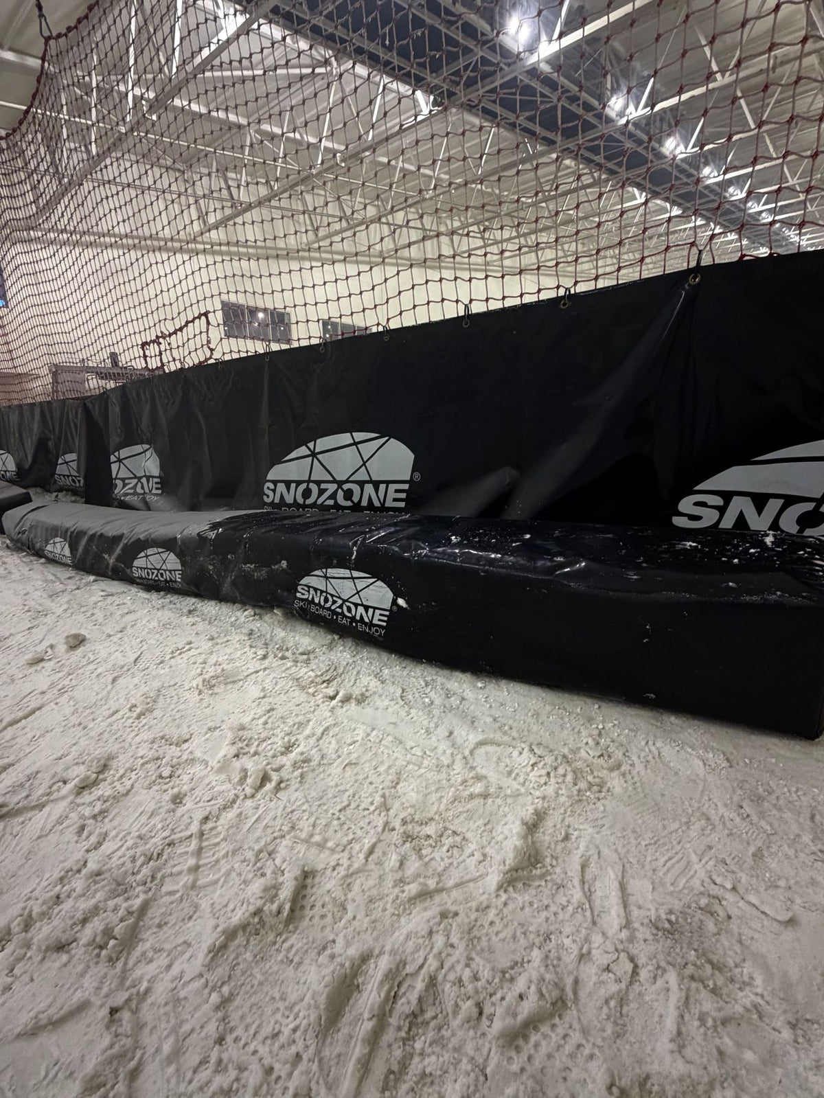 Black Snozone snowboard safety padding with white logo branding, set on a snowy indoor ski slope with a net and metal ceiling structure visible in the background.