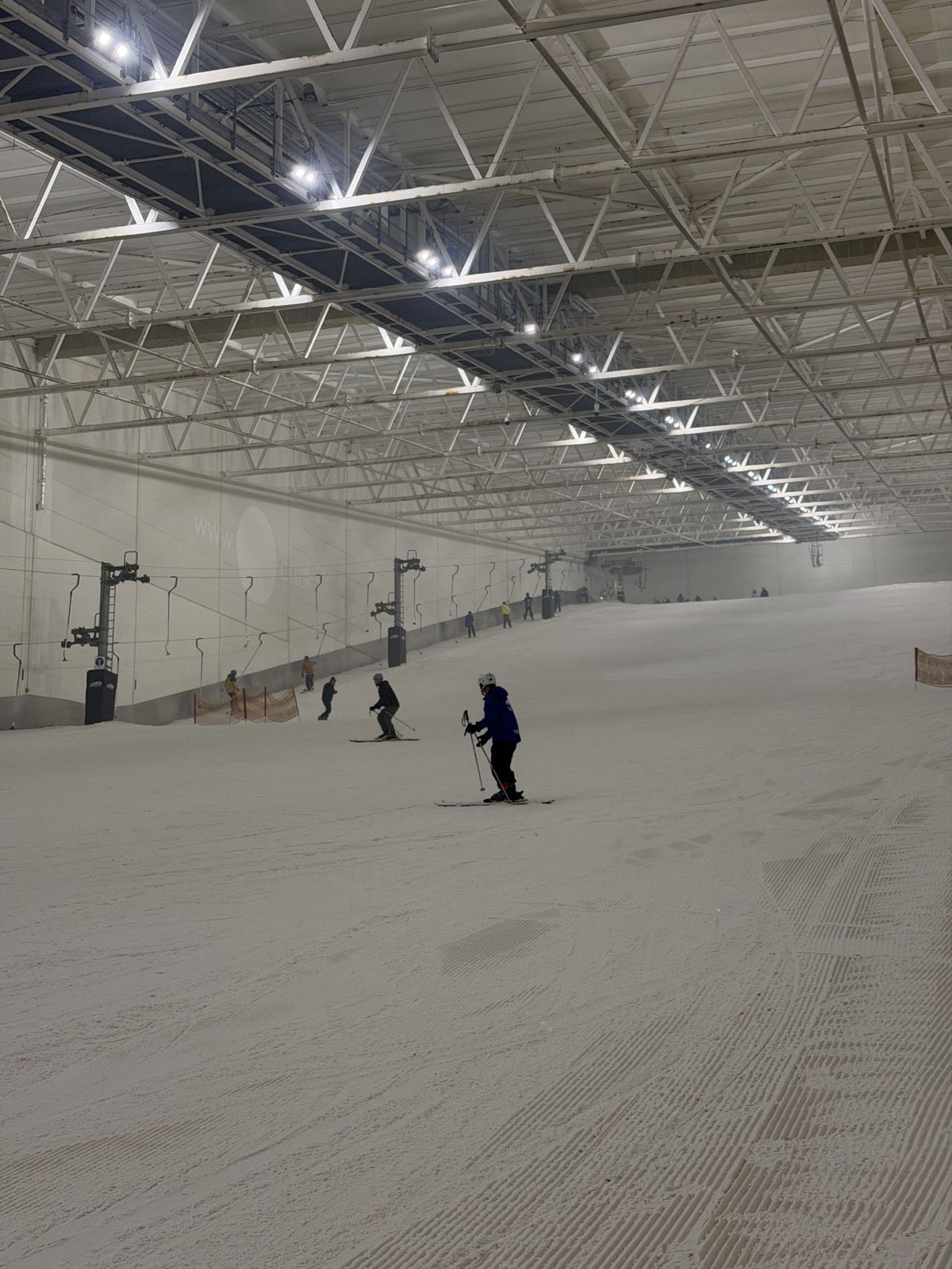 A person in dark ski gear stands on a snowy indoor ski slope under a large metal support structure with bright overhead lights. Other skiers are visible in the distance, and ski lifts are present along the left side.