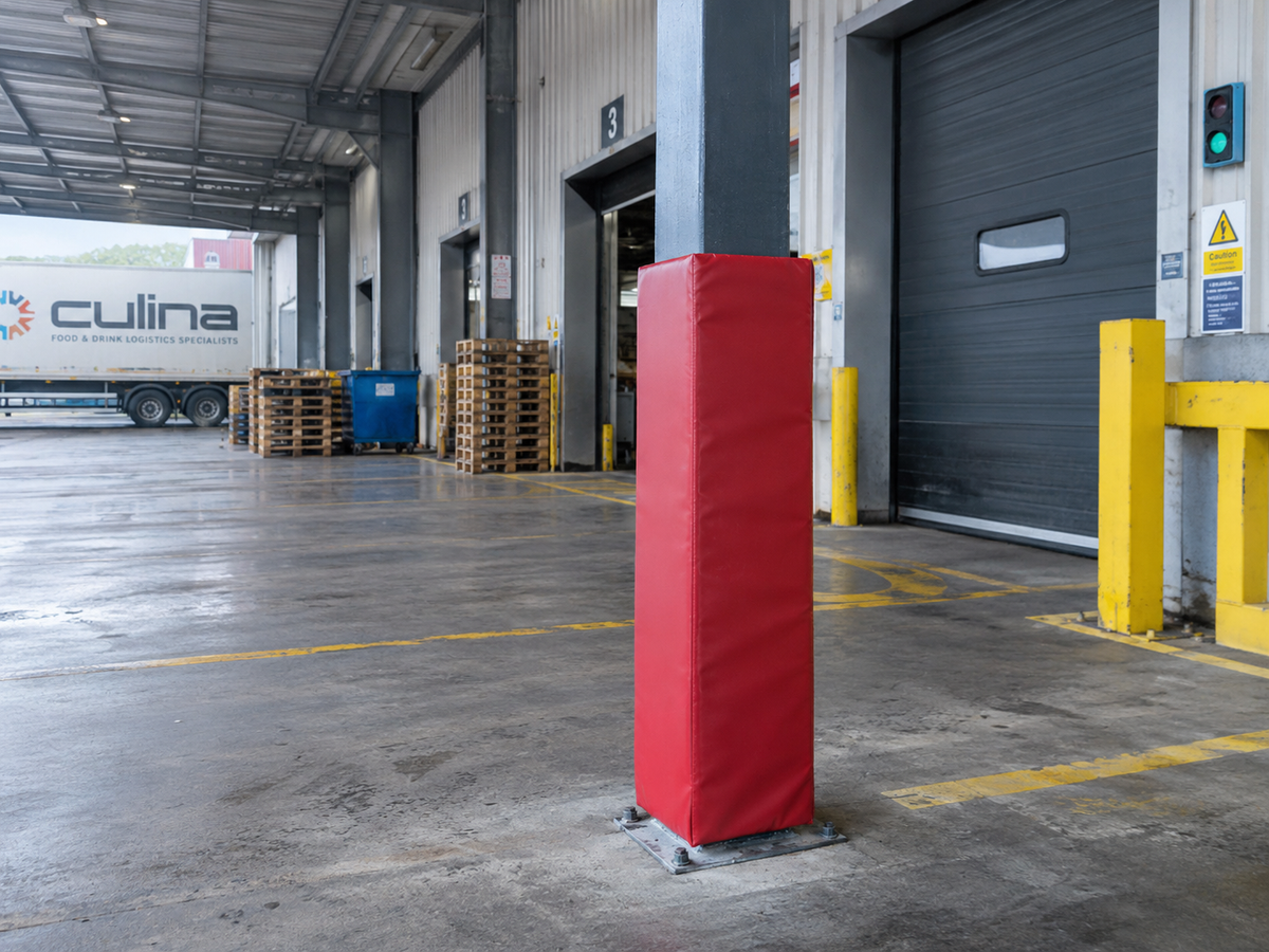 A tall red protective bollard pad with a fabric cover stands on a metal base inside a warehouse loading dock area next to a gray structural column.