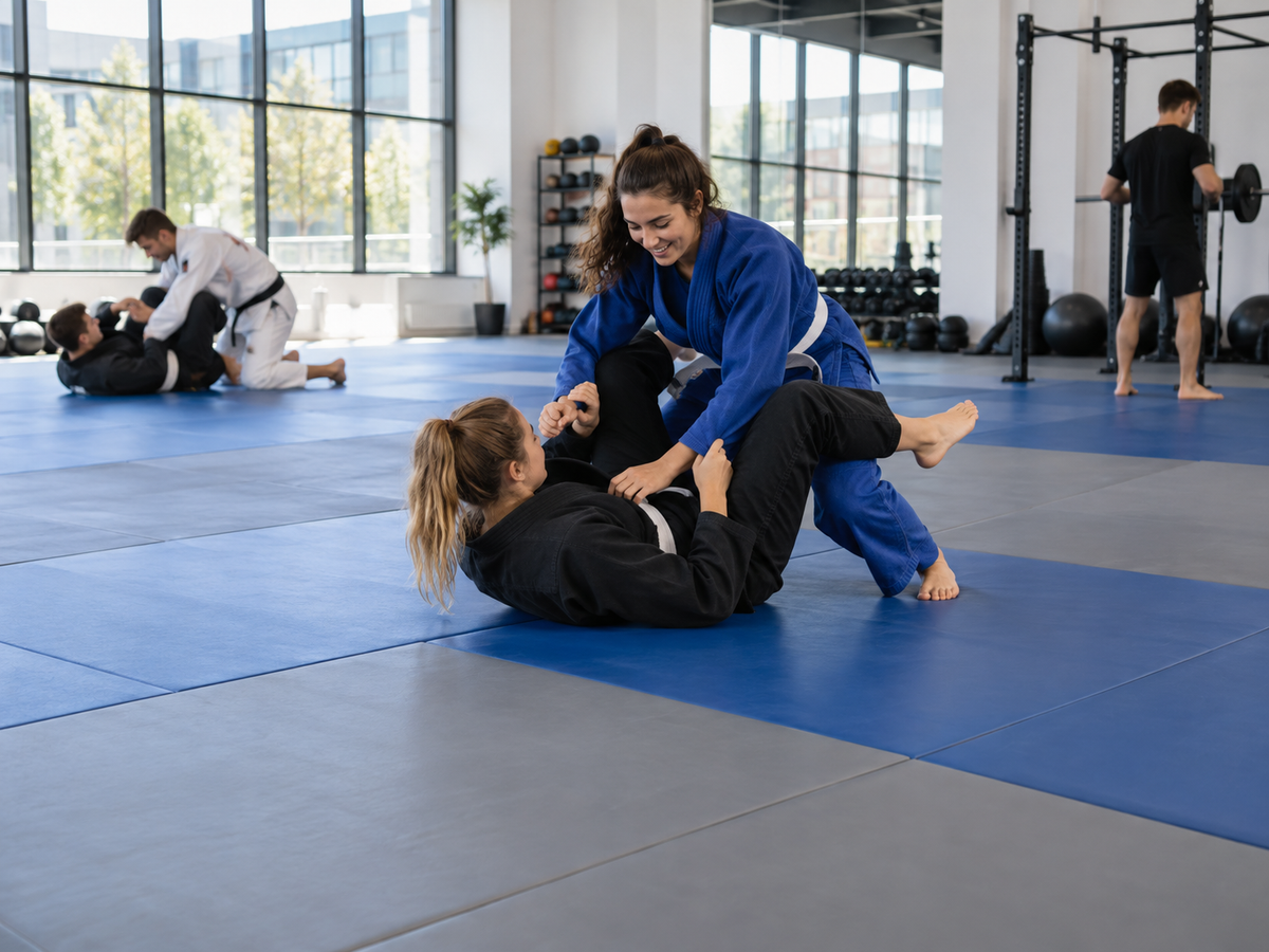 Two people practicing Brazilian jiu-jitsu on blue mats in a gym, with one person in a blue gi grappling on top of another person in a black gi.