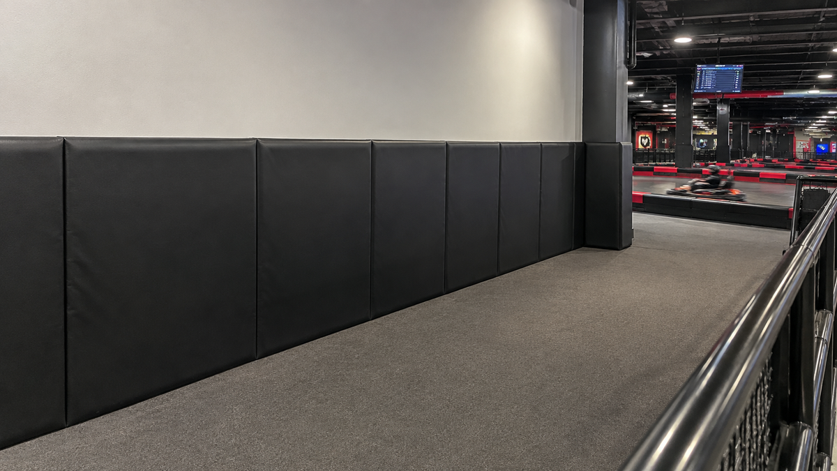 Black padded wall panels installed along a corridor inside an indoor arena or gym, with gray carpeted flooring and a metal railing in the foreground.