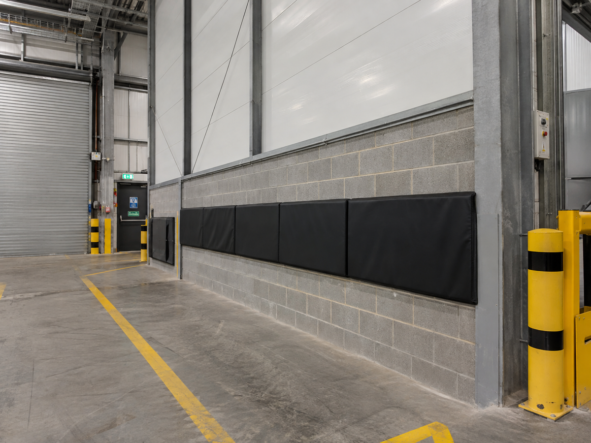 Black industrial warehouse wall protectors mounted on a gray brick wall next to a loading dock gate, with yellow safety bollards and a concrete floor in the background.