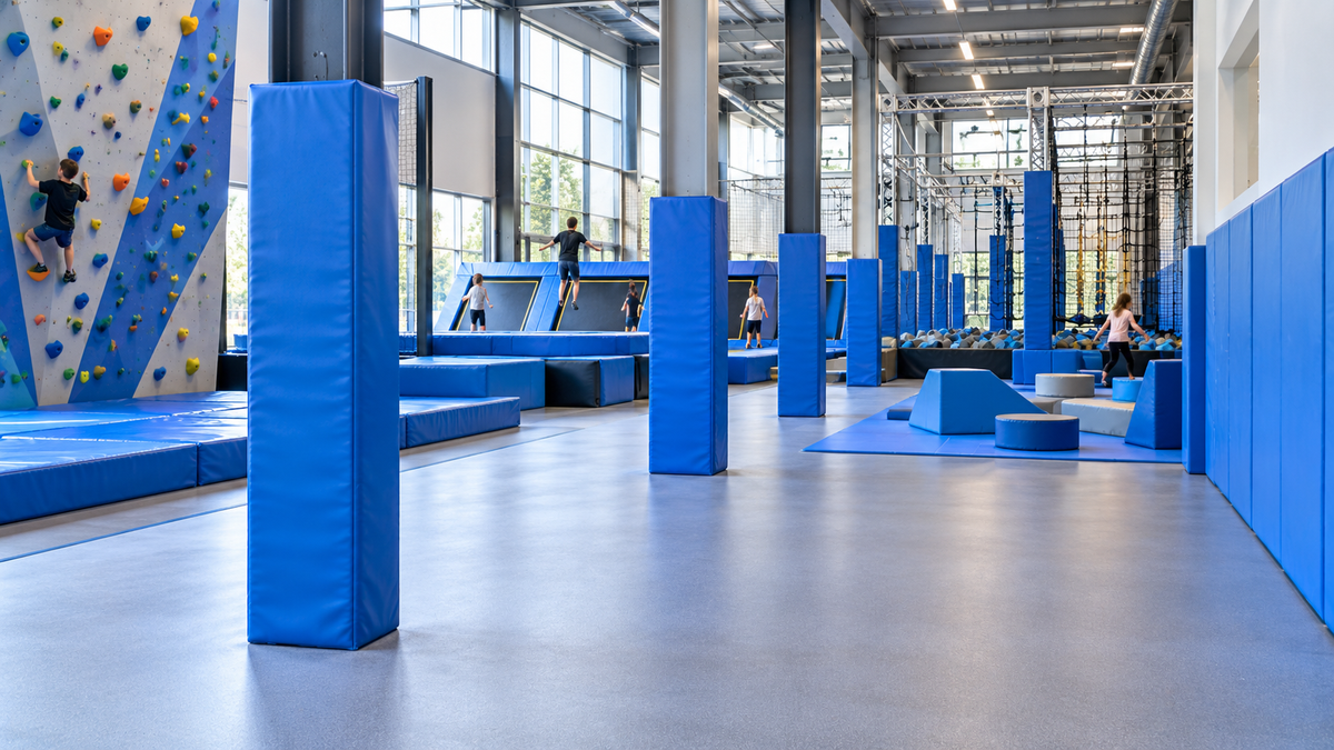 An indoor adventure playground with blue padded foam blocks, climbing walls, trampolines, and large windows in the background. Several people are visible using the facilities.