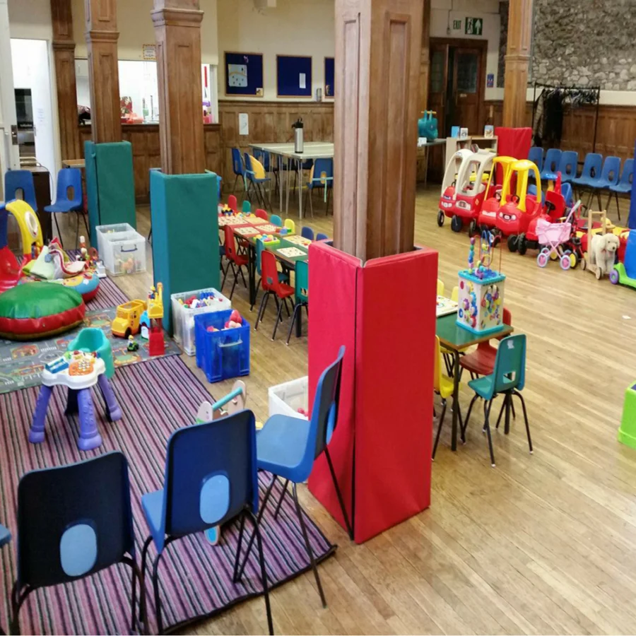 A large indoor playroom or children's activity space with wooden floors, wooden pillars, and multiple colorful children's furniture pieces and toys arranged throughout the room.